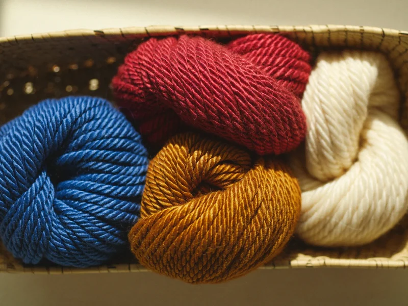 Handgesponnene Wollgarne in Krapprot, Indigo, Ocker und Naturweiß im Korb (Illustration)